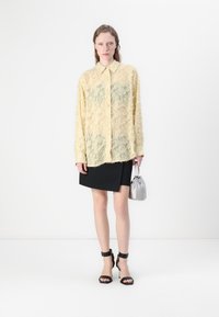Camicia gialla a maniche lunghe con texture e design trasparente; gonna mini nera; borsa argentata; sandali neri con tacco a spillo e cinturini alla caviglia.