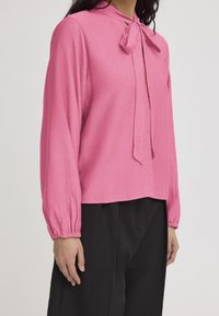 Blusa rosa a maniche lunghe con scollatura a fiocco, realizzata in tessuto strutturato e con polsini arricciati, abbinata a pantaloni neri a gamba larga.