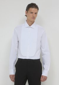 Weißes formelles Hemd mit strukturiertem Vorderteil und Knopfschluss. Lange Ärmel mit geknöpften Manschetten. Getragen mit schwarzen Hosen.