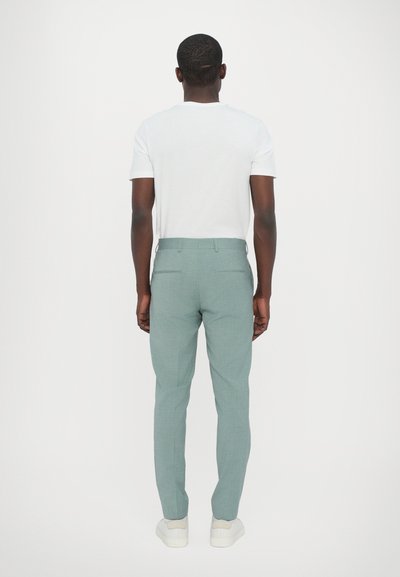 Hellgrüne, maßgeschneiderte Hose mit subtiler Textur, versehen mit Gesäßtaschen und schlanker Passform, getragen mit einem weißen T-Shirt und Sneakers.