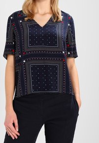 Camisa de manga corta azul marino con escote en V, que presenta un patrón geométrico de puntos rojos y estrellas blancas, hecha de una tela lisa y ligera.