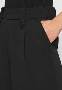 Černé kalhoty s hladkou texturou, s plochým předním dílem a dvěma bočními kapsami. Zapínají se na dva černé knoflíky v pase.