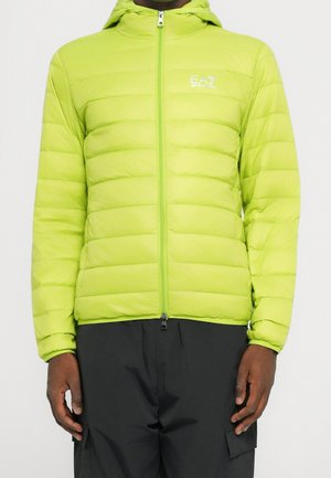 Personne portant une veste matelassée à capuche vert citron avec une fermeture éclair sur le devant et un pantalon cargo noir, debout devant un fond uni.