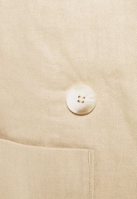 Gros plan sur un tissu beige avec un seul bouton blanc à quatre trous au-dessus du bord cousu d'une poche.