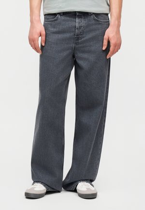 Άνδρας που φοράει φαρδιά σκούρα γκρίζα τζιν και λευκά αθλητικά παπούτσια, στέκεται με τα χέρια χαλαρά πάνω σε ένα απλό φωτεινό φόντο.