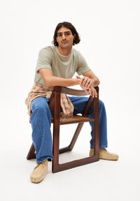 Hellgrünes T-Shirt mit kurzen Ärmeln, blaue gerade geschnittene Jeans, beige Mokassins. Sitzt auf einem Holzstuhl mit gewobenem Sitz und einem karierten Hemd, das um die Taille gebunden ist.