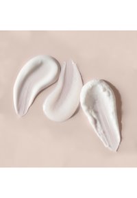 Drei sich überlappende Farbmuster eines cremigen kosmetischen Produkts in unterschiedlichen Formen, mit weißen und hell-lila Farbtönen, vor einem sanften beigen Hintergrund.