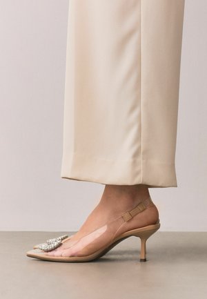 Pied portant un escarpin slingback beige à talon kitten avec côtés en PVC clair et détail nœud en cristal, associé à un pantalon crème à jambes larges.