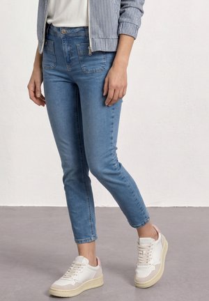Personne portant un jean slim bleu, des baskets blanches avec des accents beige, un haut blanc et une veste rayée, debout sur un sol gris.