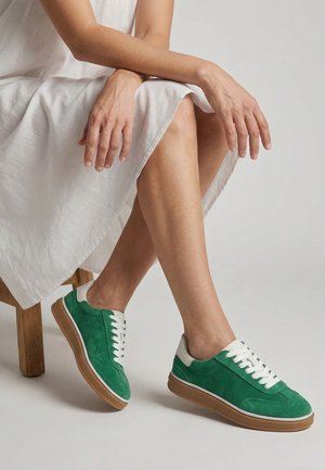 Person iført hvid kjole og grønne ruskindssneakers med gummisåler, siddende på en træskammel, hænder hvilende på korslagte ben.