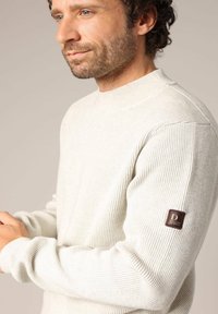 Lichtbeige ribbeltrui met hoge hals, voorzien van een bruine leren patch op de mouw met reliëf logo-detail. Zachte textuur.