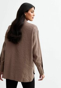 Lange mouwen, lichtbruine blouse van een gestructureerde stof, met drop shoulders en zijslitsen, gezien van achteren.
