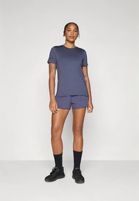 Mulher de pé a usar uma t-shirt atlética azul-marinho e calções, meias pretas e sapatilhas pretas, contra um fundo claro e liso.