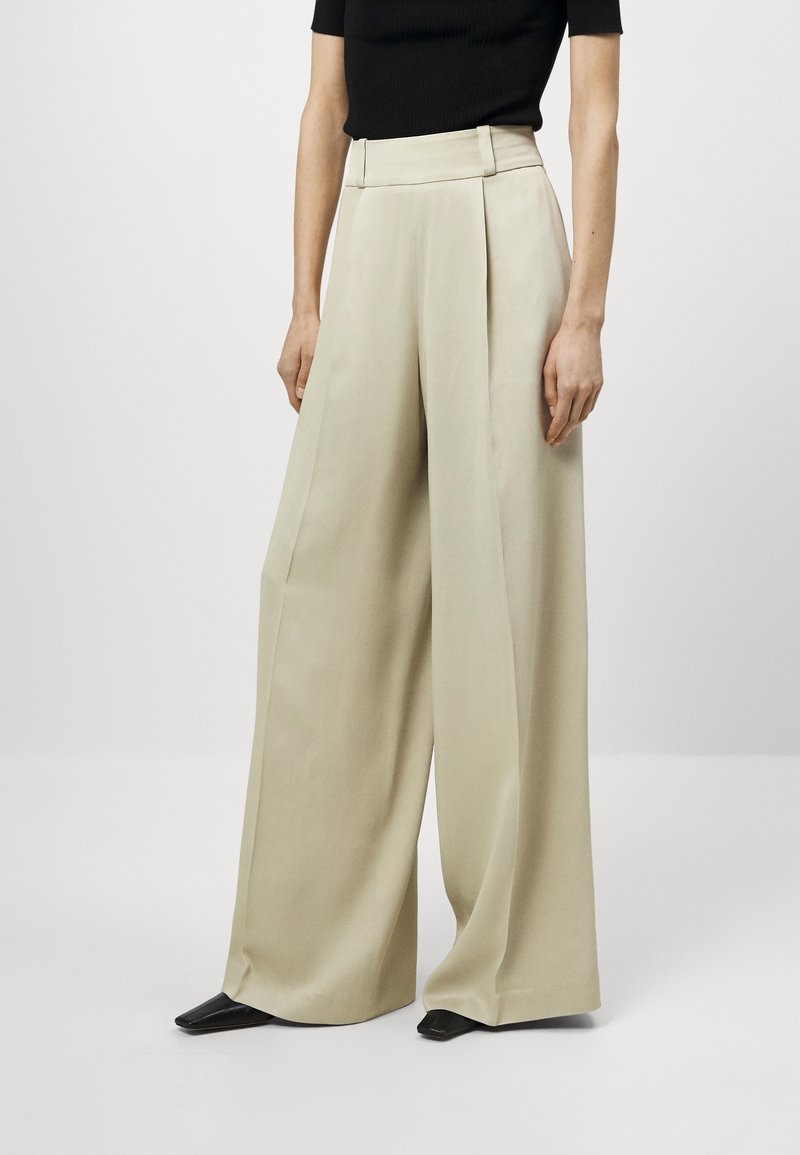 Crème brede pantalon met plooien, hoge taille en gladde stof; gecombineerd met een aansluitend zwart topje en puntige zwarte schoenen.