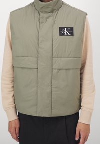 Prošívaná vesta olivově zelené barvy s vysokým límcem, dvěma předními kapsami a černou nášivkou s logem. Nošena přes světle béžové tričko s dlouhým rukávem.