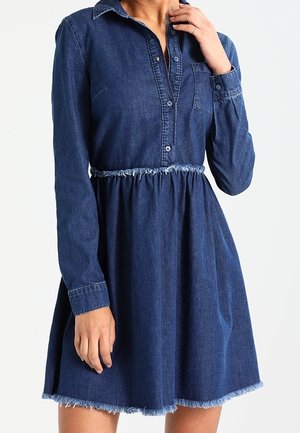 Robe en denim bleu foncé à manches longues, boutonnée à l'avant, avec une poche poitrine et des bords effilochés à la taille et à l'ourlet, portée par une personne.
