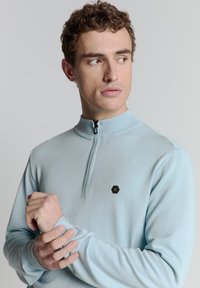 Lichtblauwe zip-up trui met een opstaande kraag, ribgebreide manchetten en een klein zwart hexagonaal logo op de borst. Glad, gebreid textuur.