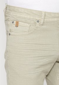 Pantalon en jean beige avec poche avant et coutures visibles, porté avec une chemise blanche rentrée à la taille.