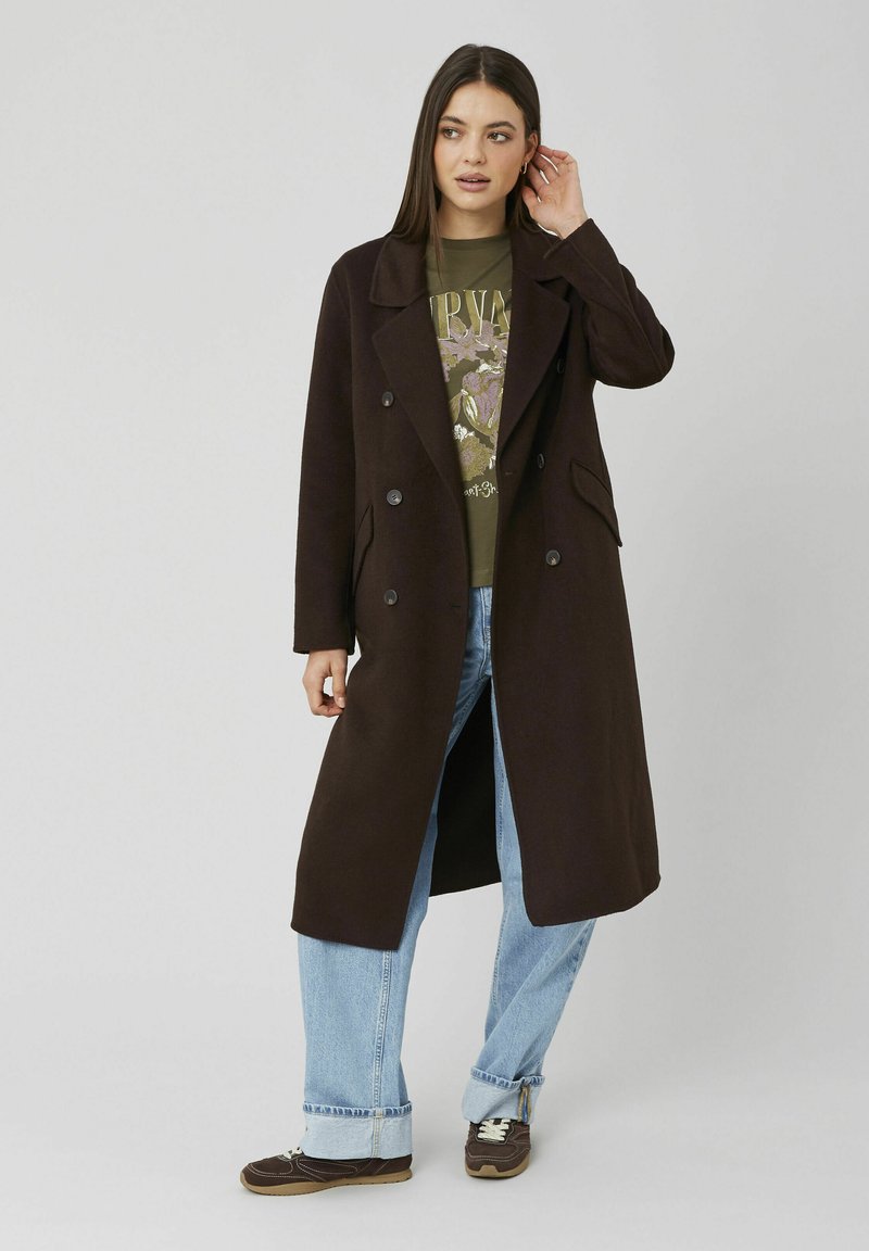 Manteau long à double boutonnage de couleur brun foncé avec des revers à pointe, des poches latérales et une coupe décontractée, porté sur un t-shirt vert à motif et un jean bleu.