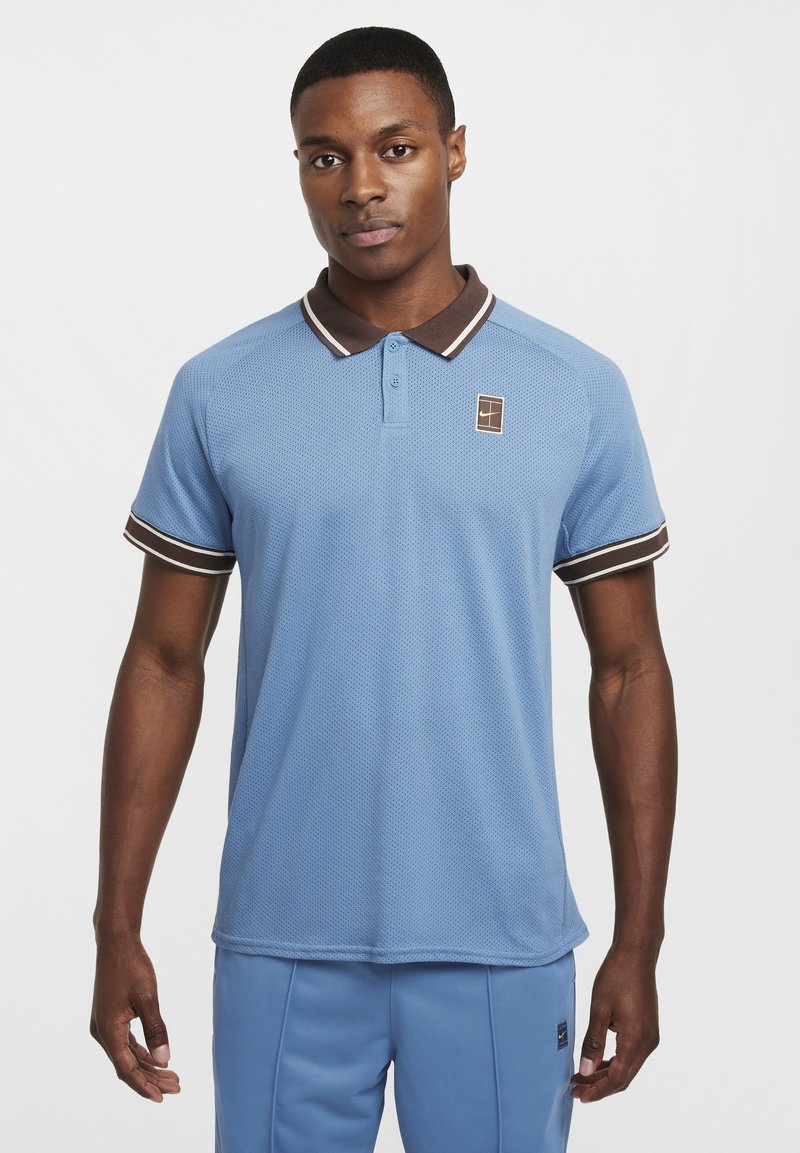 Lys blå polo-skjorte med teksturert mesh-stoff, brun krage og stripete detaljer på ermene. Har en liten logo-patch på brystet.