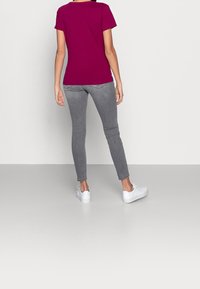 Magenta T-Shirt mit kurzen Ärmeln kombiniert mit grauen Slim-Fit-Jeans und weißen Sneakern, von hinten vor einem grauen Hintergrund betrachtet.