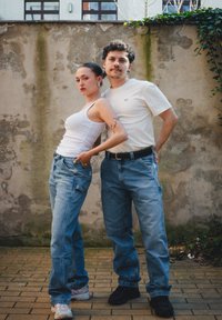Due persone posano di fronte a un muro testurizzato. Una indossa una canotta bianca e pantaloni baggy blu chiaro; l'altra una maglietta crema e jeans scuri.