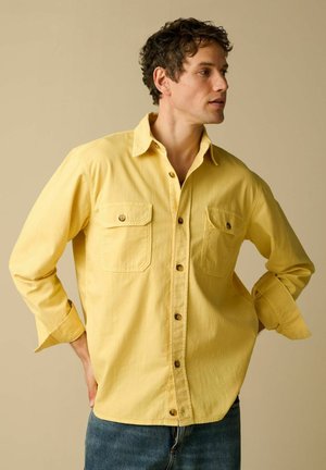 Homme portant une chemise jaune boutonnée avec deux poches poitrine, les mains sur les hanches, regardant vers la droite sur un fond beige uni.