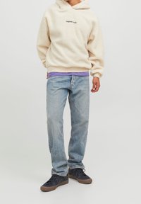 Felpa in pile color crema con cappuccio e coulisse, indossata sopra una maglietta lavanda, abbinata a jeans in denim azzurri chiari e sneakers scure.
