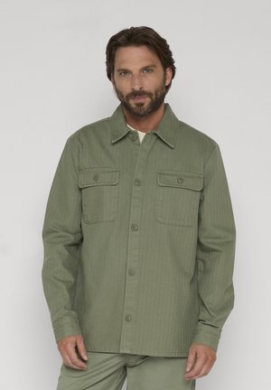 Homme barbu portant une veste olive verte boutonnée avec deux poches poitrine, debout devant un fond clair uni.