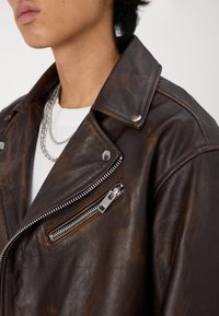 Giacca in pelle marrone con una texture liscia, colletto a pressione e accessori in argento. Caratterizzata da una tasca con zip frontale e una collana a catena stratificata.