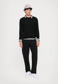 Schwarzer Strickpullover über einem karierten Hemd, kombiniert mit schwarzen Hosen und weißen Sneakers. Das Model trägt eine beige Mütze.