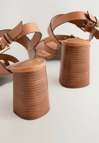Sandales à talons en cuir marron avec de larges sangles, présentant un talon en bois texturé et rayé ainsi que des accents en métal doré.