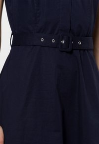 Robe marine avec une taille cintrée et une ceinture en tissu, ornée de ferrures argentées, et une texture en coton lisse. Comprend des détails de perforations sur la ceinture.