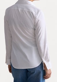 Camisa blanca de manga larga con cuello, con una textura suave y puños con un solo botón, combinada con jeans de mezclilla azul.