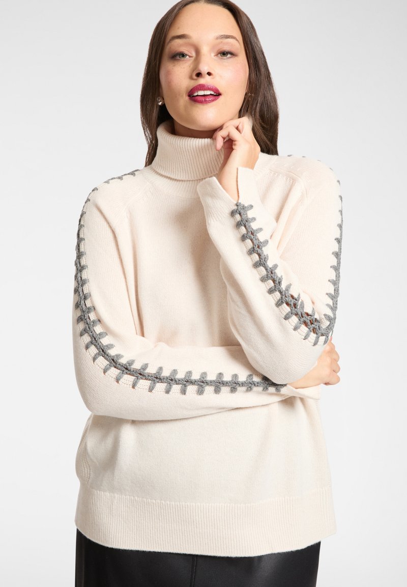 Jasnobeżowy sweter dzianinowy z wysokim kołnierzem, szarymi plecionymi akcentami na rękawach oraz ściągaczami na mankietach i dole.
