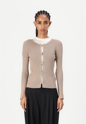 Vaale beige ribbed-cardigan, jossa on pitkät hihat, napitus, hienovarainen pinta ja halkiot helmassa. Päällä valkoinen, kuvioitu paita ja mustat housut.