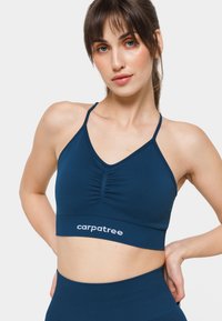 Tumši zilo sporta krūšturis ar regulējamām siksnām, V veida dekoltē, tekstūrēta auduma un plandā siena priekšā. Ar baltu logotipu.