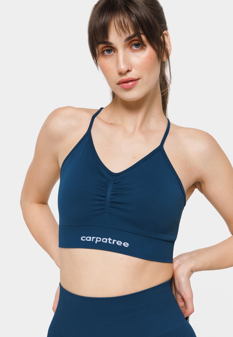 Tumši zilo sporta krūšturis ar regulējamām siksnām, V veida dekoltē, tekstūrēta auduma un plandā siena priekšā. Ar baltu logotipu.