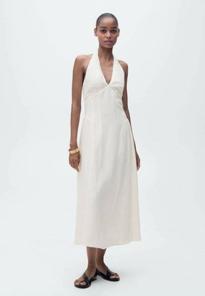 Un vestito halter di lunghezza al ginocchio, di colore crema, con una profonda scollatura a V e una silhouette fluente, realizzato in tessuto leggero, abbinato a sandali neri.