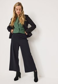 Svart kavaj över en grön blus, kombinerad med högmidjade, vida svarta byxor och svarta ankellånga stövlar. Släta texturer, avslappnad look.