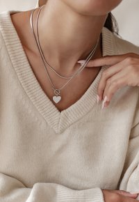 Zilveren gelaagde colliers met een gladde hartpendant, gedragen over een lichtbeige gebreide trui met een diepe V-hals.