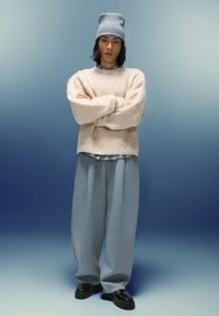 Jasnofioletowy, puszysty sweter, szare, oversize'owe dresy, czarne buty oraz jasnoniebieska, dziana czapka z teksturowanym wzorem, na tle gradientu.