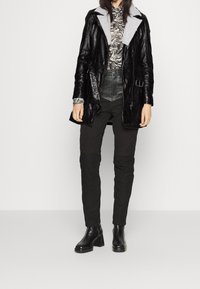Abrigo de cuero negro con cuello de piel sintética gris, combinado con una blusa estampada y pantalones negros ajustados. Botines negros gruesos completan el look.