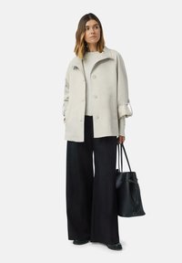Manteau trench beige clair avec un large col, fermeture à boutons et manches retroussées ; associé à un pantalon noir à larges jambes et un sac noir.
