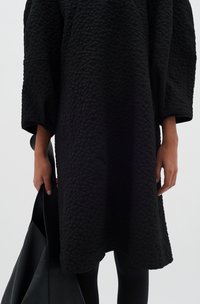 Gepatchtes schwarzes Kleid mit weiten Ärmeln und lockerer Passform, kombiniert mit einer schwarzen Ledertasche, die eine glatte Textur und ein elegantes Design aufweist.