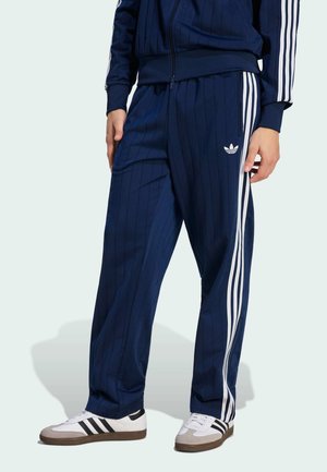 Osoba nosząca granatowe spodnie dresowe i kurtkę Adidas z białymi paskami, w połączeniu z białymi tenisówkami z czarnymi paskami.