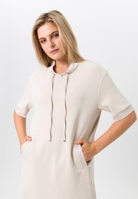 Robe pull beige à manches courtes, col à cordon de serrage et poches latérales. Tissu doux avec un léger éclat et détails ajustés.
