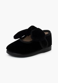 Zapatilla de terciopelo negro con punta redondeada, que presenta suaves orejas de conejo exageradas y un borde cosido, descansando sobre una suela de goma plana.