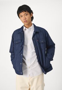 Giacca leggera color blu navy con bottoni a pressione, due tasche frontali e una texture liscia, indossata sopra una camicia bianca a righe.