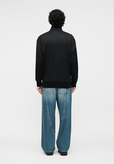 Άνδρας με σγουρά μαλλιά που φοράει μαύρο μακρυμάνικο τοπ και φαρδιά μπλε τζιν παντελόνια, στέκεται με την πλάτη προς το φακό μπροστά σε απλό λευκό φόντο.